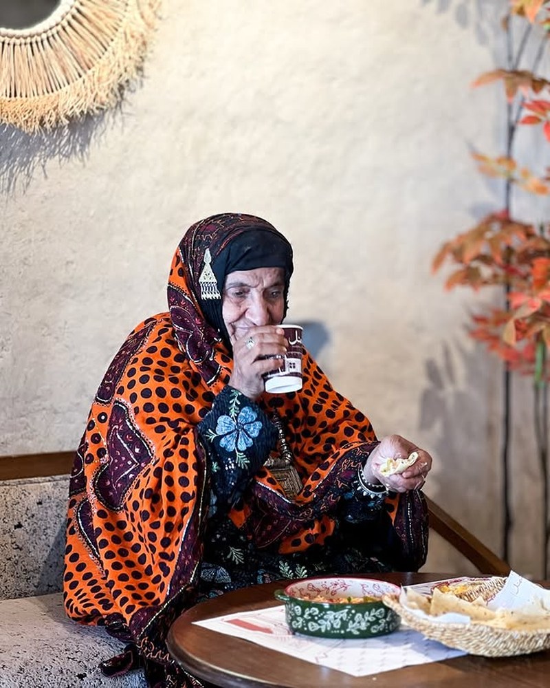 سيدة عُمانية مسنّة بالزي التقليدي تحتسي الشاي مع خبزٍ في أجواء شَمْل الحجرية ببهلا